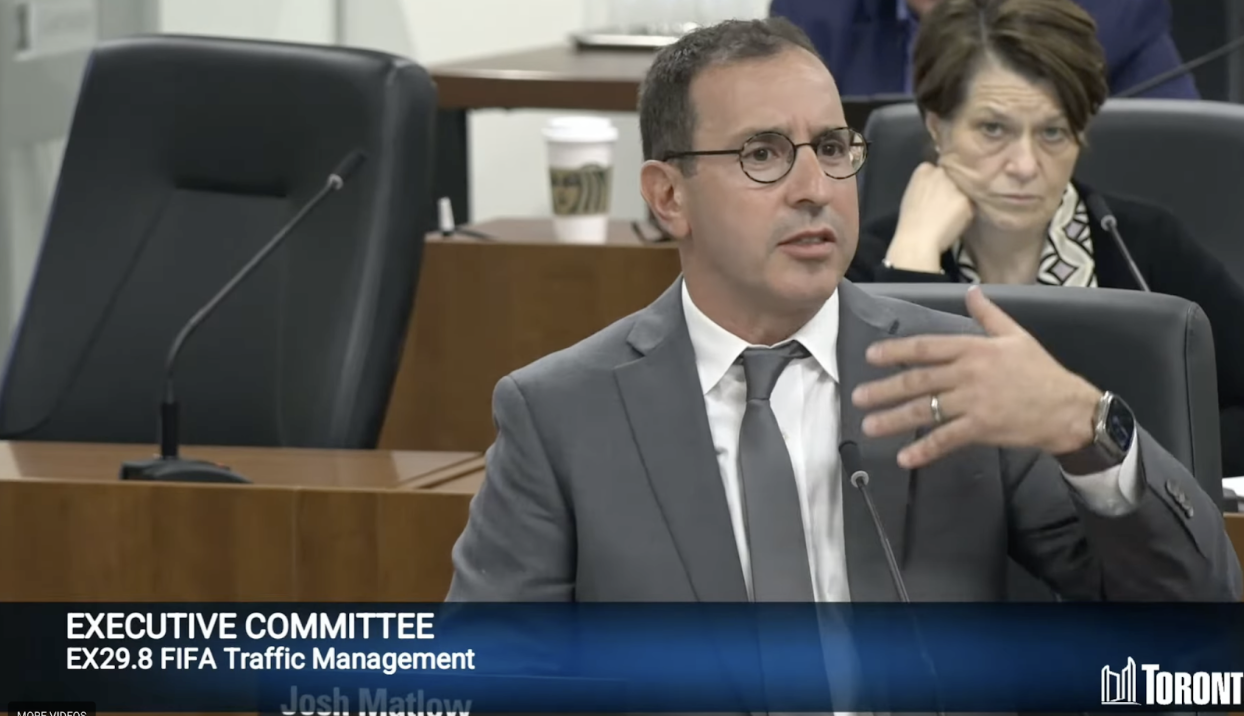 A man dressed in a suit and tie, sitting in front of a microphone, speaking.