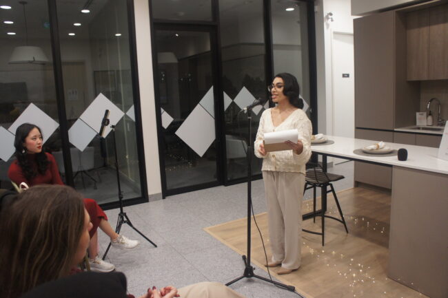 A woman stands speaking into a microphone, while holding papers, to an audience seated in a room with glass walls and a kitchen setup.