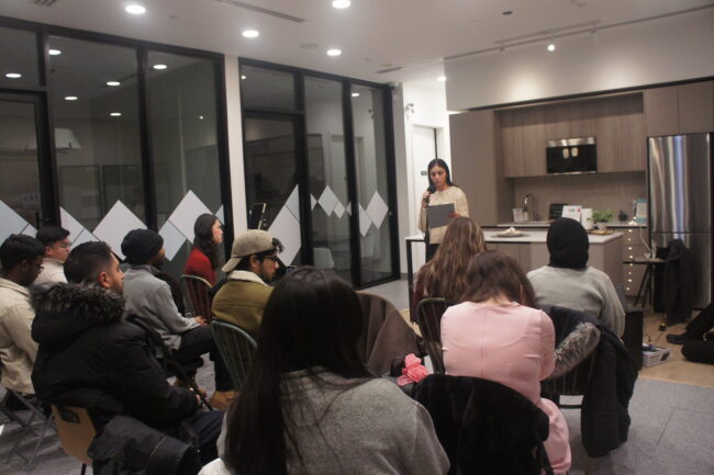 A woman stands speaking into a microphone to an audience seated in a room with glass walls and a kitchen setup