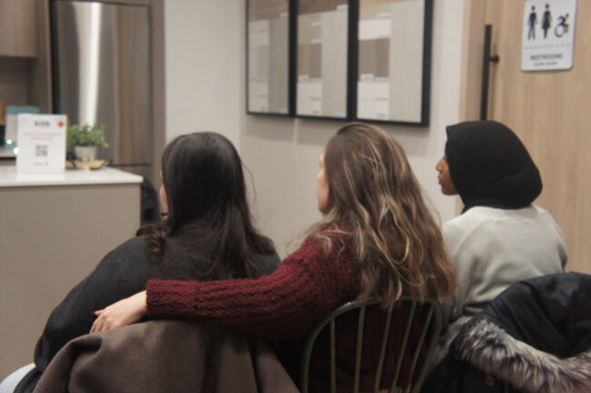 Three people sitting in a room, with their backs to the camera. The one in the middle has their arm around the one to their left.