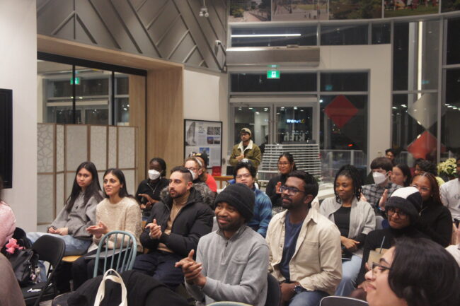 A diverse audience in a room listen and clap.