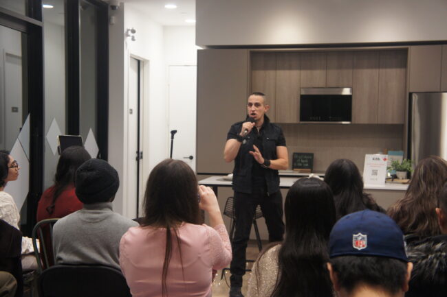 A person in a black shirt speaks into a microphone in front of an audience in a room with glass walls and a kitchen setup. An audience member is recording with their device.