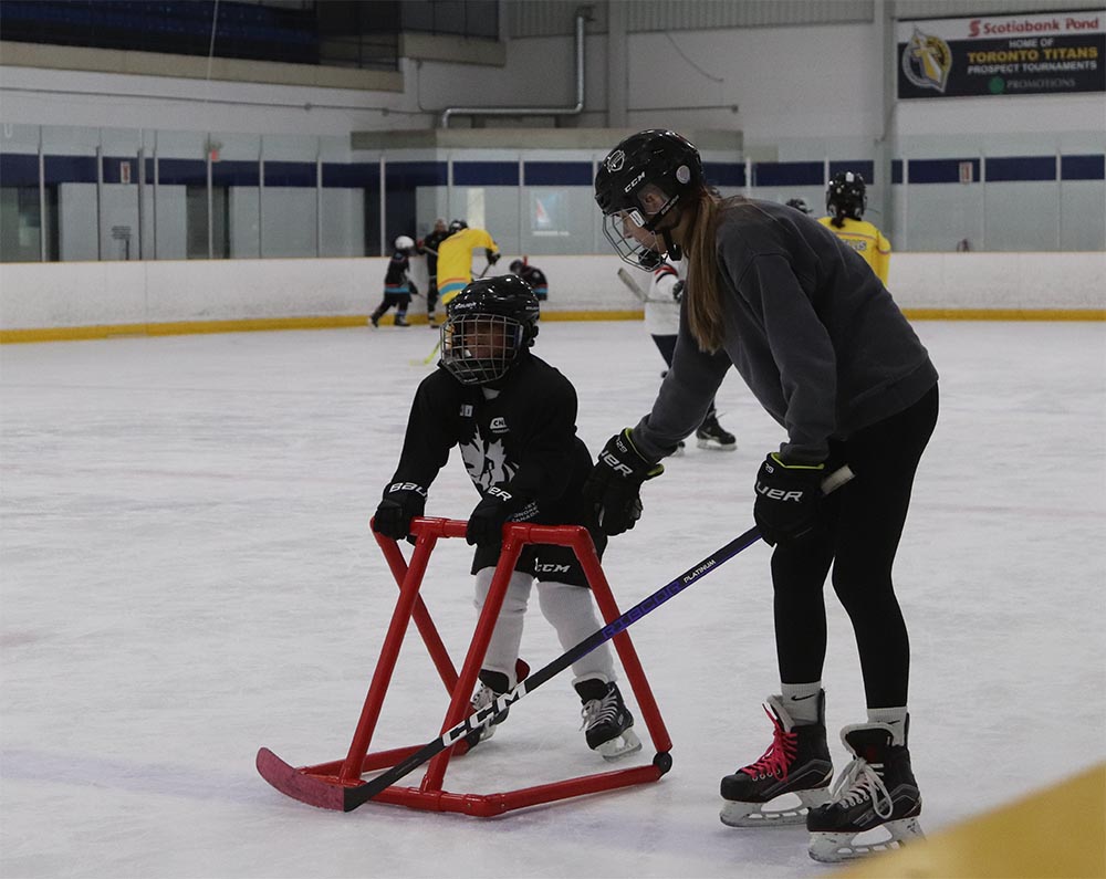 A girl guides a partially sighted boy who is learning how to skate on the ice. He holds onto a skating aid trainer.
