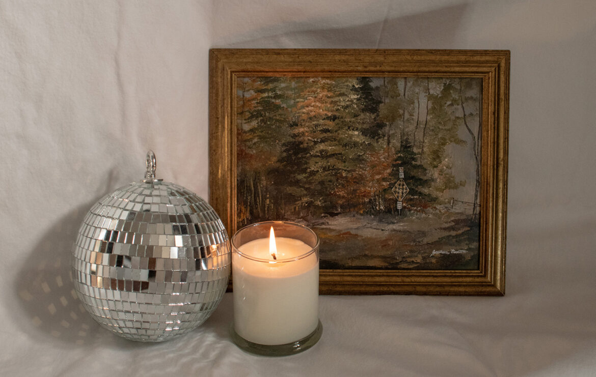 A framed landscape painting with a disco ball and lit candle in the centre of a white background.