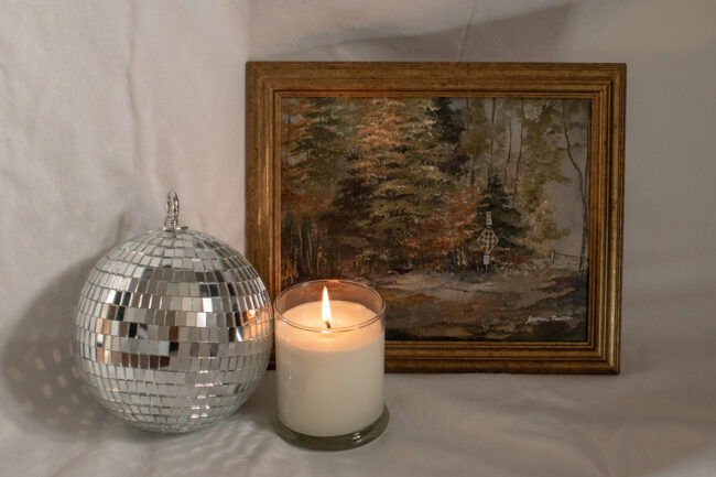 A framed landscape painting with a disco ball and lit candle in the centre of a white background.