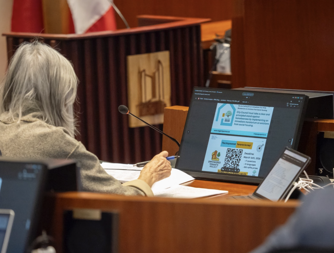 A person with grey hair sits at a desk in a conference room, viewing a presentation on a large monitor.