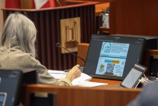 A person with grey hair sits at a desk in a conference room, viewing a presentation on a large monitor.