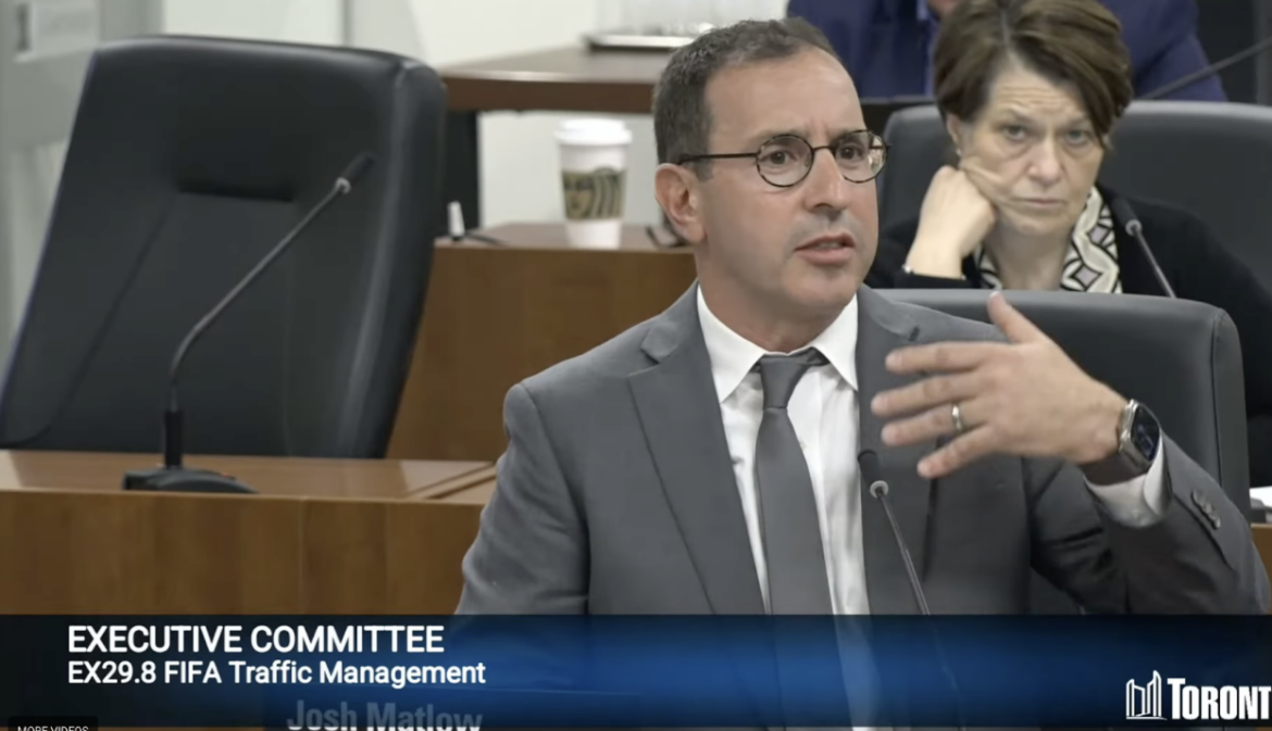 A man dressed in a suit and tie, sitting in front of a microphone, speaking.
