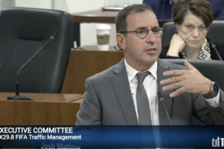 A man dressed in a suit and tie, sitting in front of a microphone, speaking.