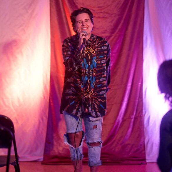 A person smiling while holding a microphone in front of a pink-purple fabric backdrop.
