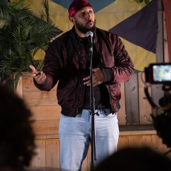 Persons speaking into a microphone with a Jamaican flag behind them. A video recording device from an audience member is partially visible.