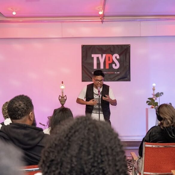 Person speaking into a microphone on a stage in front of an audience, while holding their phone. A black banner behind them reads "TYPS"