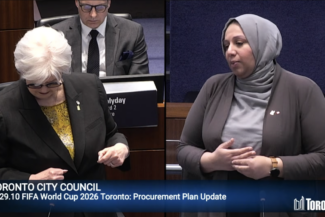 Two people speaking at a Toronto City Council meeting. One wears a black suit and glasses, and the other wears a grey hijab