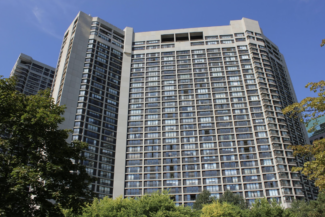 Tall residential high-rise with numerous windows under a clear blue sky, surrounded by green trees